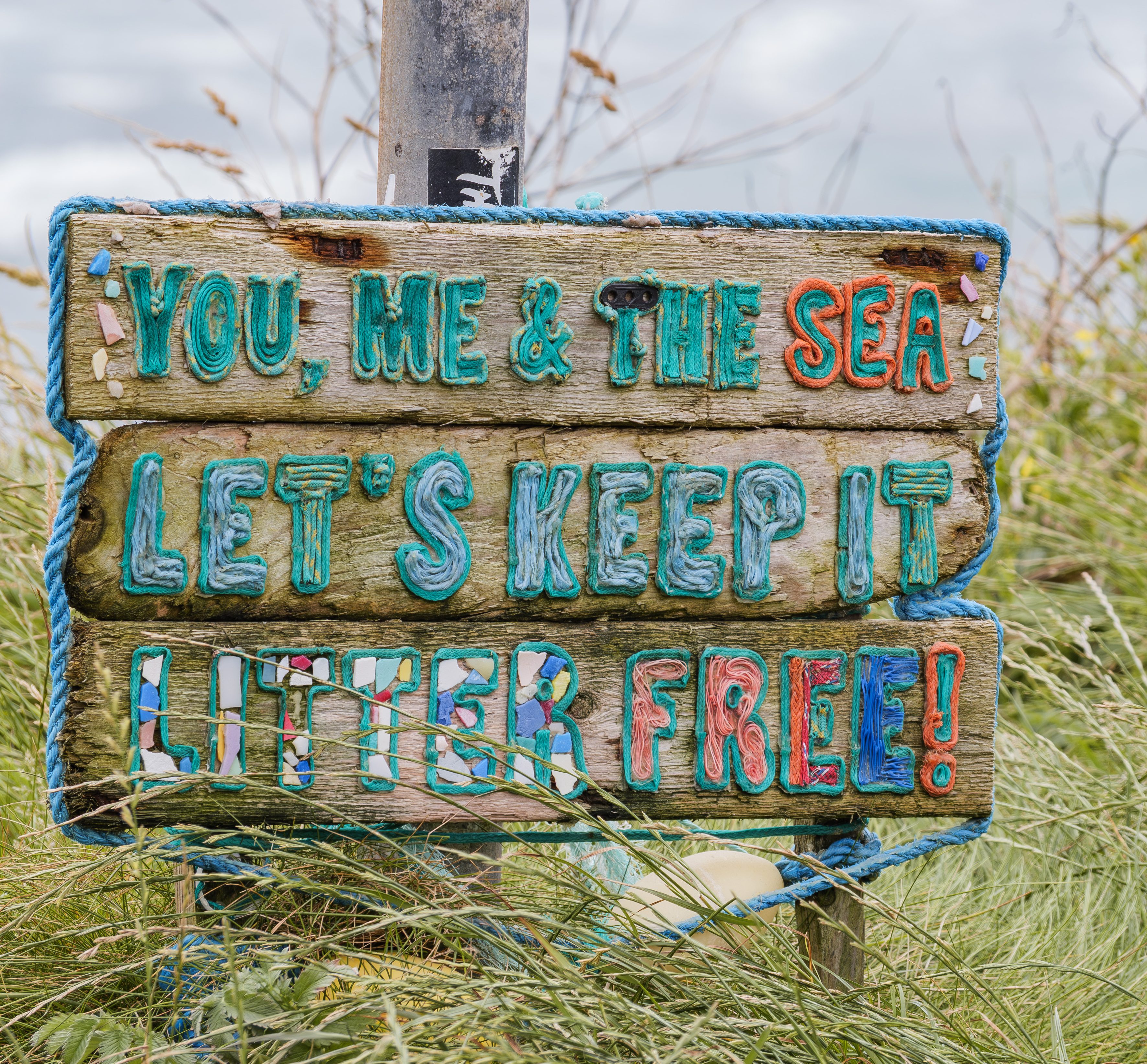 Ein bunt bemaltes Holzschild aus Treibholz mit der Aufschrift You me and the sea im Dünengras an der irischen Küste.