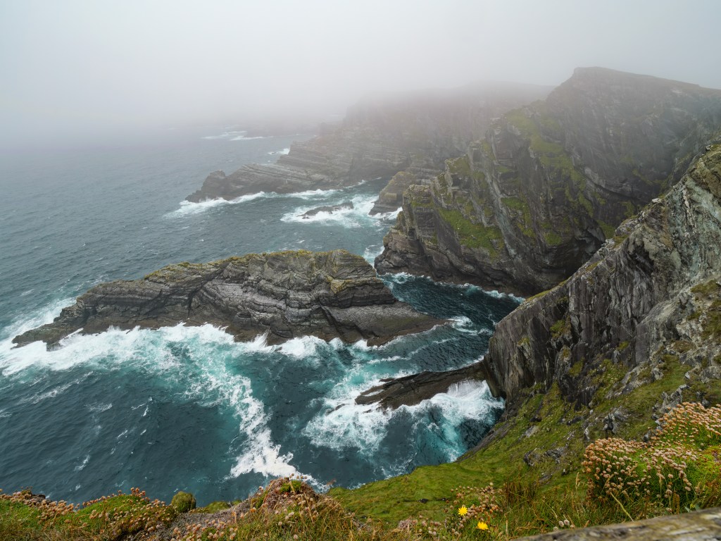 Blick von oben auf die rauen Kerry Cliffs mit im Nebel verschwindenden Felsspitzen und türkisfarbener Brandung.