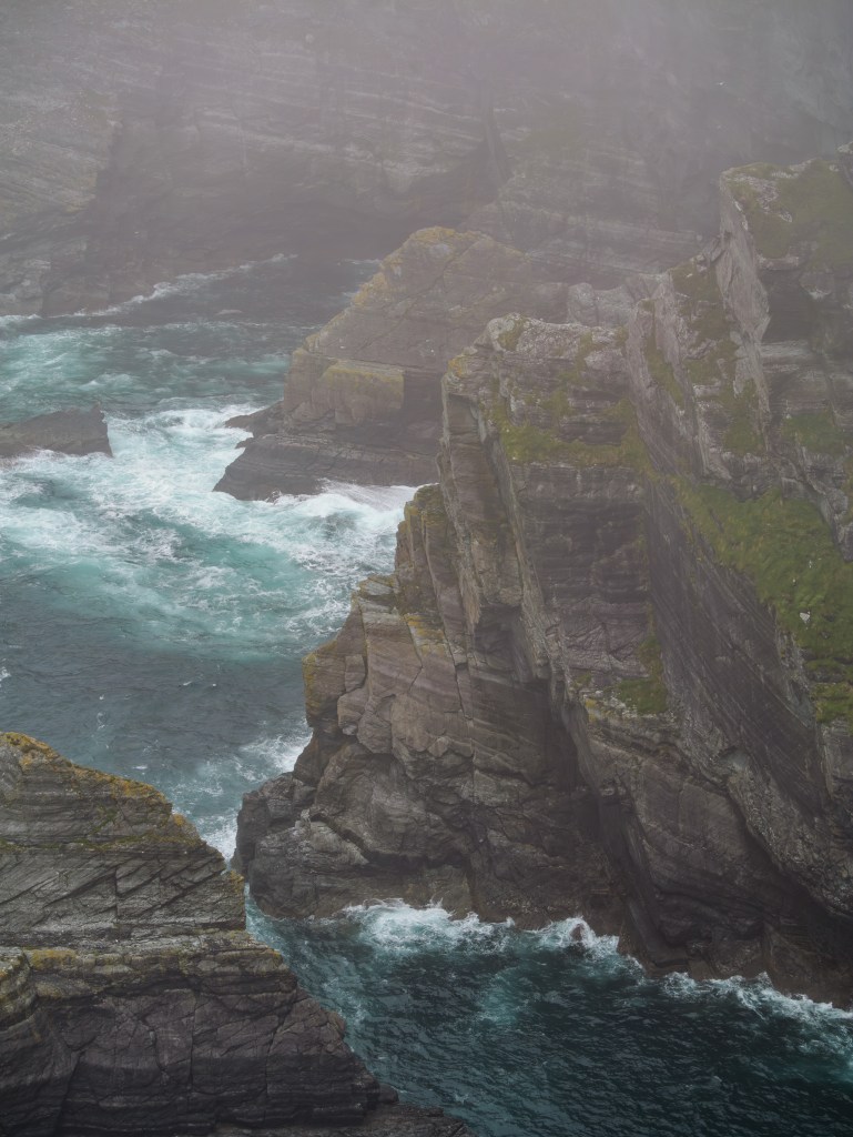 Nahaufnahme der schroffen, dunklen Felswände der Kerry Cliffs inmitten einer dichten Wolken- und Nebelsuppe.