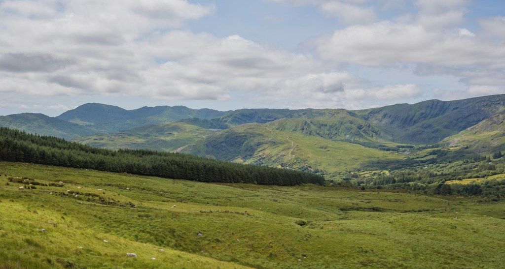 Querformat-Landschaftsaufnahme von einer schmalen Serpentinenstraße durch die grünen Berge und Täler am Caha Pass in Irland bei wechselhaftem Wolkenlicht.